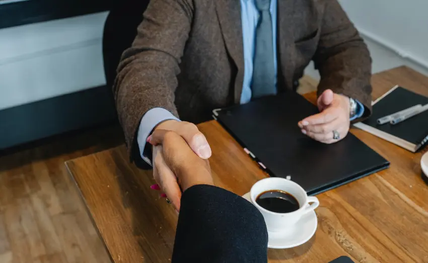 Two people shaking hands over a table with coffee.